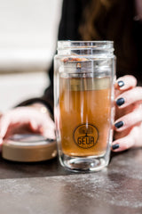 A close-up of a hand holding a clear double-walled glass mug from the "Teeflasche Bambus Edition 450ml" collection by GEVA Vital, filled with tea. The premium-quality mug features a visible tea infuser inside and an exterior logo that reads "GEVA Vital." The person's other hand is reaching for the auslaufsicher (leak-proof) lid, which is lying on the table.