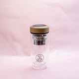 A clear, double-walled glass Teeflasche from the Teeflasche Bambus Edition 450ml collection by GEVA Vital, featuring a beige lid and metal infuser basket. The bottle, exhibiting Premium-Qualität and the "GEVA" logo on its side, stands elegantly against a soft pink background.