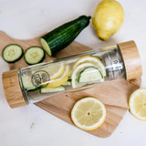 A GEVA Vital Teeflasche Bambus Edition 450ml, filled with lemon and cucumber slices, is placed on a wooden cutting board. The premium-quality bottle features bamboo lids at both ends to ensure it is leak-proof. Next to the bottle, additional lemon and cucumber slices accompany a whole lemon and a partially sliced cucumber.