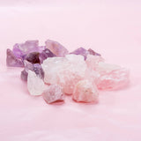 A collection of Edelsteine from GEVA Vital's Edelsteinwasser Basis-Set Amethyst + Bergkristall + Rosenquarz, featuring light pink, white, and purple stones such as amethyst, displayed on a pastel pink surface. The Natursteine are irregularly shaped, with their raw, natural texture visible.
