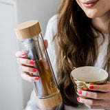 A woman with long brown hair and pink nail polish smiles slightly, holding a GEVA Vital Teeflasche Bambus Edition 450ml – Dein 2in1-Teegenuss für unterwegs in one hand and a decorative teacup in the other.