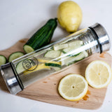 The GEVA Vital Teeflasche Stahl Edition 450ml, filled with cucumber and lemon slices, rests on a wooden cutting board beside a whole cucumber, whole lemon, and fresh lemon slices on a white surface.