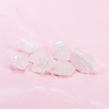 Several rough, translucent white quartz crystals from the GEVA Vital Edelsteinwasser Basis-Set Amethyst + Bergkristall + Rosenquarz are scattered on a soft pink background. The crystals vary in size and shape, displaying natural, jagged edges and imperfections. The pink background contrasts with the clear, frosty appearance of the quartz crystals.