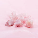 A set of raw Edelsteinwasser Basis-Set Amethyst, Bergkristall, and Rosenquarz stones from GEVA Vital arranged on a light pink background. The stones vary in size and shape, showcasing their natural, unpolished form and soft pink hues of the Rosenquarz beautifully complemented by the deep purple tones of the Amethyst and the clear clarity of the Bergkristall.